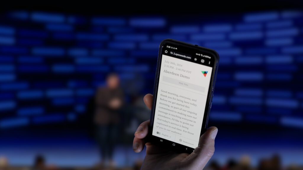 Photo of a hand holding a phone up at church, reading live captions during a sermon.