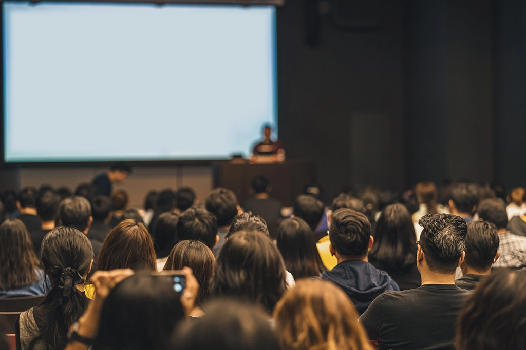 Large audience attending a university orientation or lecture with a speaker presenting on stage in front of a projection screen.