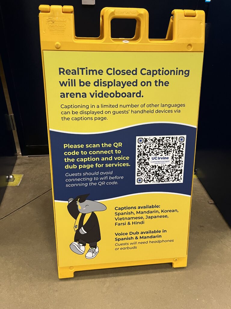 A-frame sign at a UC Irvine commencement venue directing guests to real-time closed captioning, multilingual captions, and live voice dubs via QR code, with captions also displayed on the arena video board.